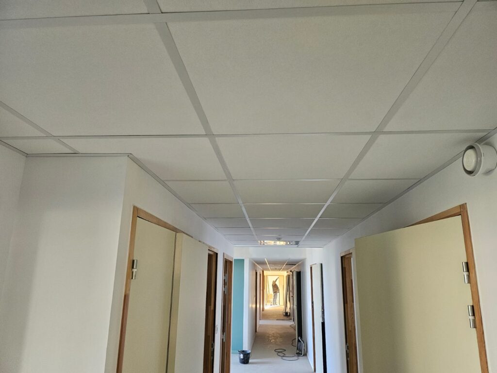 Couloir en chantier avec plafond suspendu, murs blancs et encadrements de portes en bois. Travaux de rénovation.