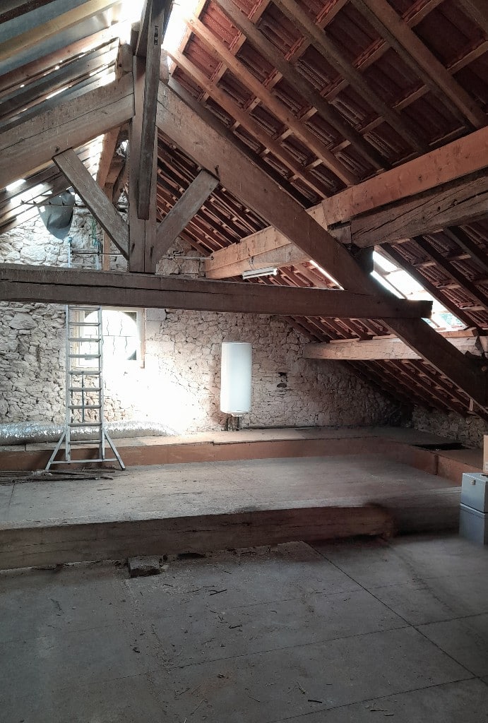 Combles en rénovation avec charpente bois apparente, murs en pierre, échelle et chauffe-eau blanc sous toiture.