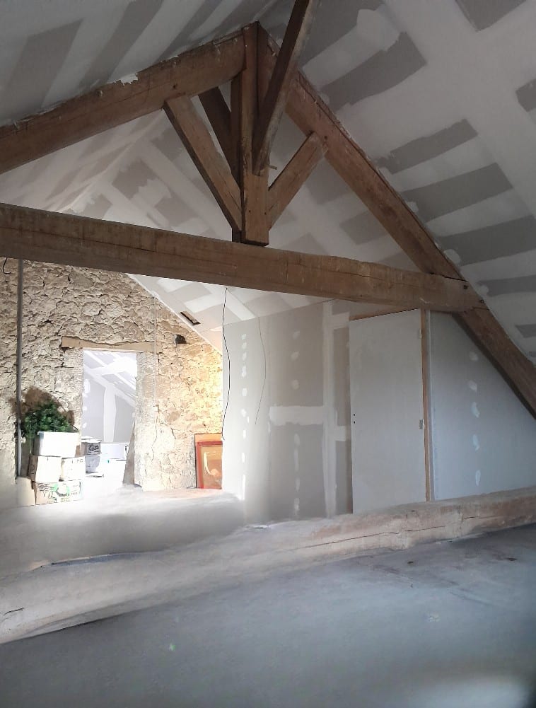 Aménagement de combles avec charpente bois apparente, mur en pierre et plaques de plâtre en cours.