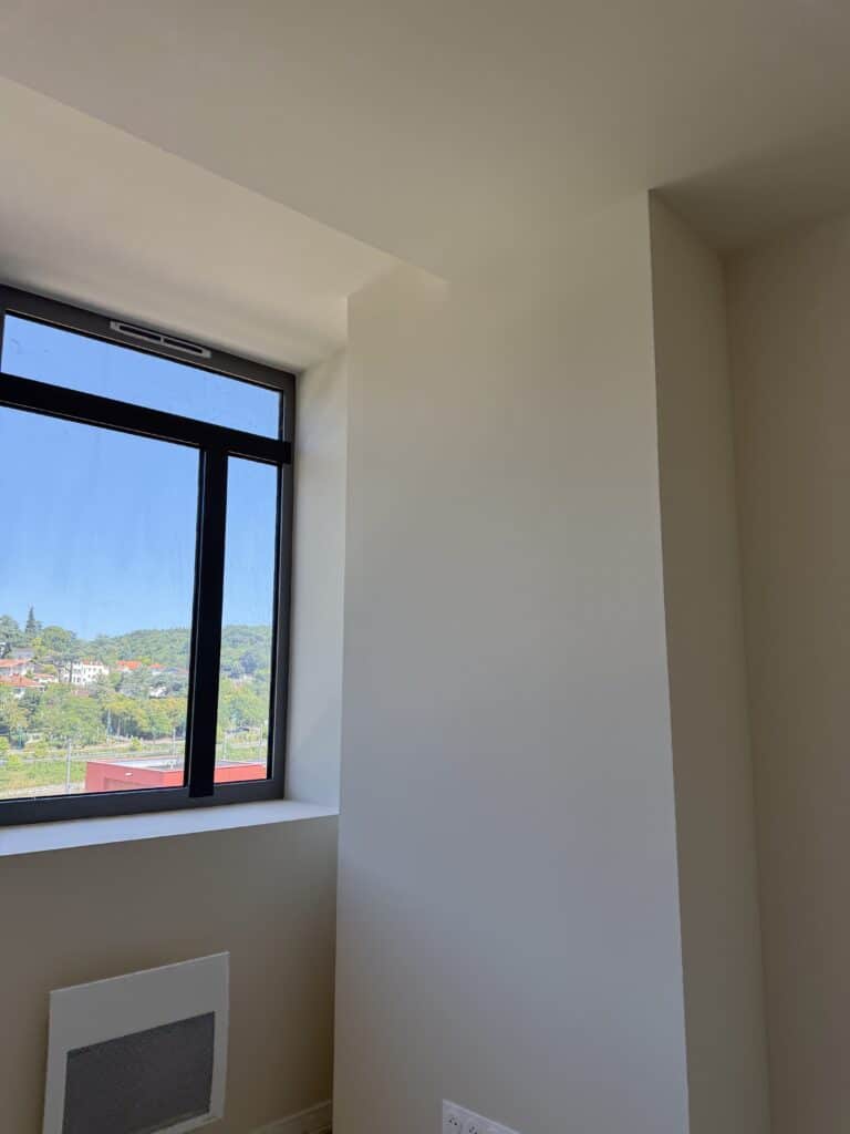 Fenêtre à cadre noir vue sur collines, intérieur moderne aux murs blancs et radiateur.