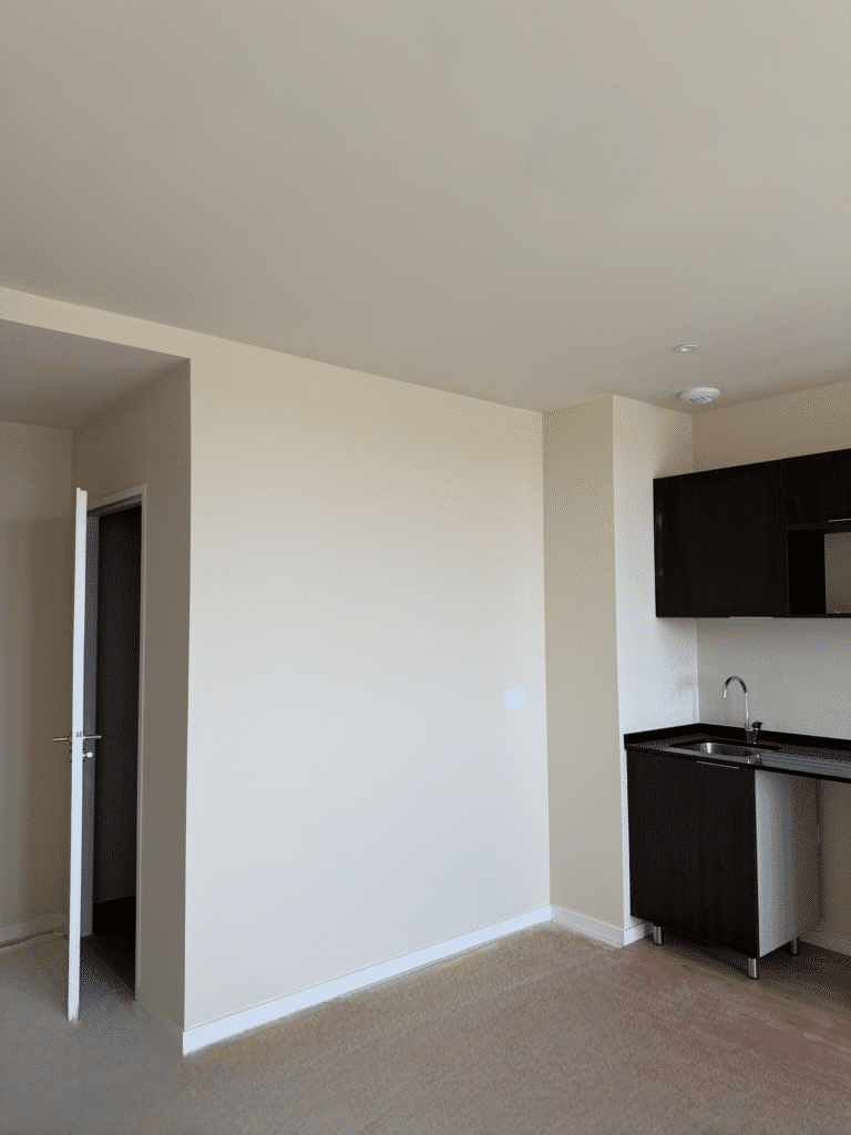 Studio moderne avec murs beiges, porte blanche ouverte et kitchenette sombre avec évier.